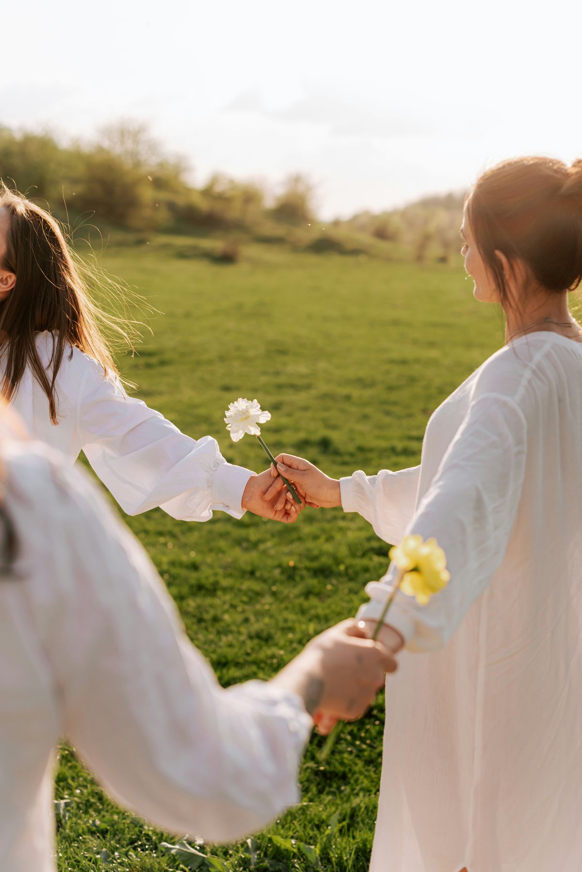 Women in white standing in a meadow at golden hour, passing flowers hand to hand